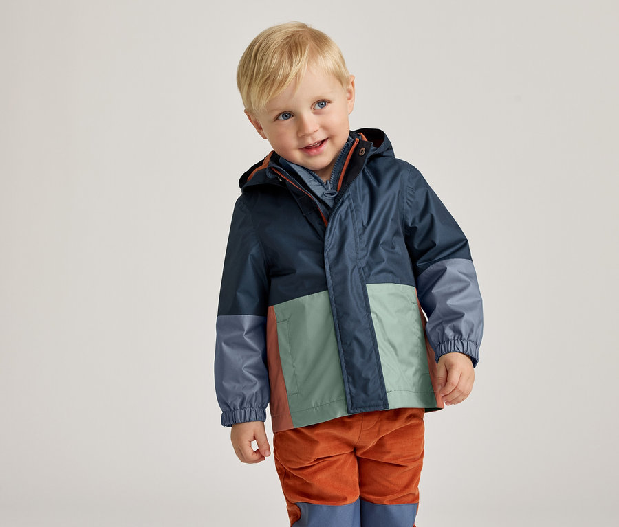 Un garçon blond souriant porte un imperméable bleu marine à blocs de couleurs et un pantalon en velours côtelé orange.