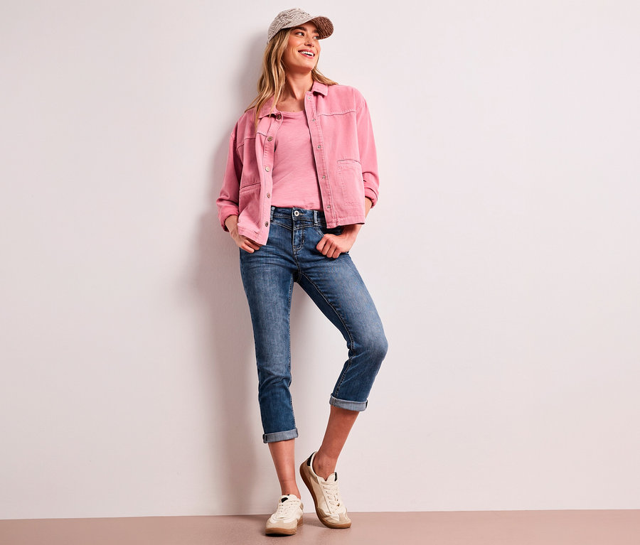 Femme souriante portant une casquette à motifs, une veste rose, un t-shirt rose et un jean 7/8 Street One « Jane » bleu avec ourlets retroussés, appuyée contre un mur blanc.