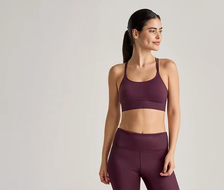 Une femme portant des vêtements de sport couleur prune, comprenant un soutien-gorge de sport à bretelles fines et un legging, pose devant un fond clair.