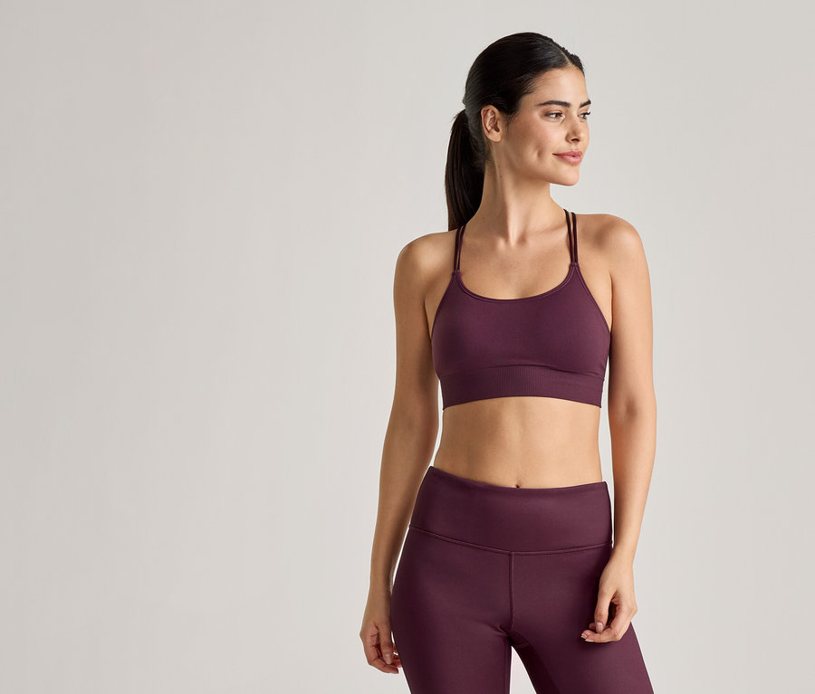 Une femme portant des vêtements de sport couleur prune, comprenant un soutien-gorge de sport à bretelles fines et un legging, pose devant un fond clair.