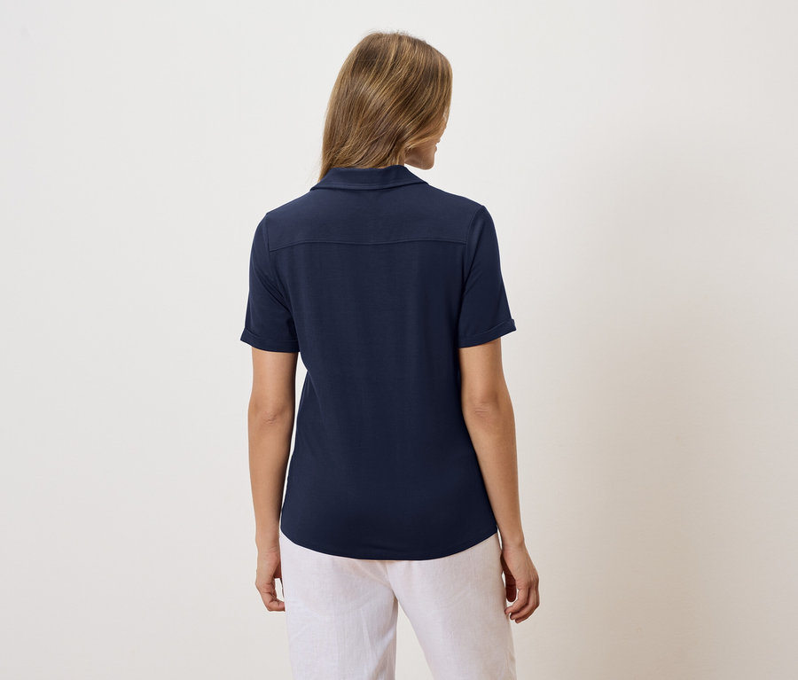 Vue arrière d'une femme blonde portant un polo bleu marine et un pantalon blanc.