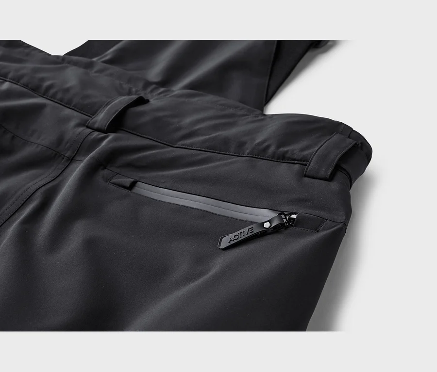 Gros plan d'un pantalon technique noir, montrant une poche zippée avec une tirette noire 'ACTIVE'. La taille avec les passants de ceinture est visible au-dessus de la poche.