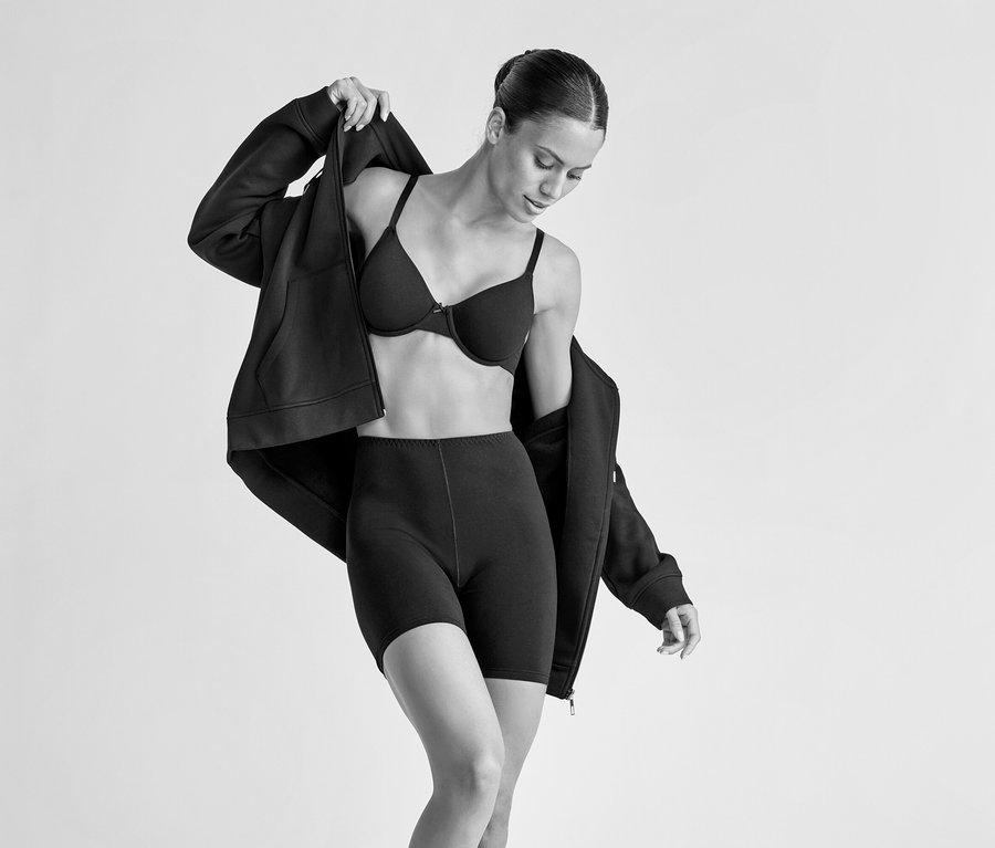 Une femme portant un soutien-gorge à armatures noir et un short noir enfile un sweat à capuche zippé noir. Elle regarde vers le bas en tirant le sweat sur ses épaules.