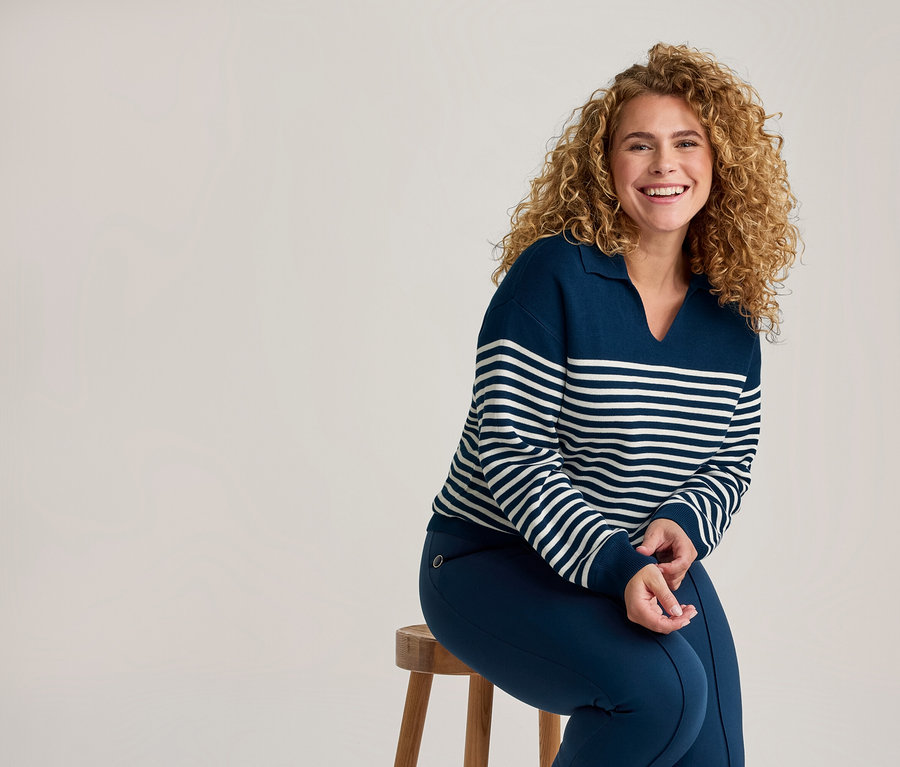 Femme souriante aux cheveux bouclés portant un pull rayé bleu marine et blanc et un pantalon bleu marine, assise sur un tabouret en bois.