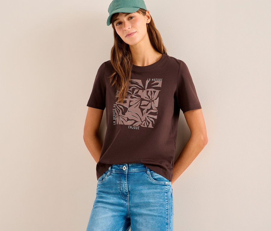 Femme portant un t-shirt à motif CECIL marron avec un motif floral, un jean bleu clair et une casquette verte. Elle est appuyée contre un mur clair.