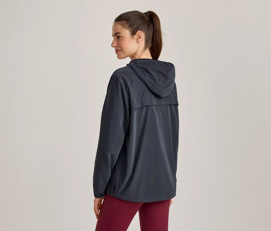 Femme debout, dos au spectateur, la tête légèrement tournée vers la droite. Elle porte une veste à capuche gris foncé et un legging rouge foncé.