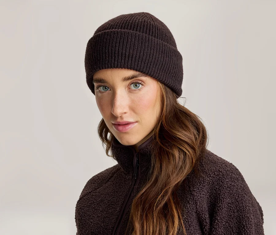 Femme aux longs cheveux bruns, portant un bonnet côtelé tricoté marron foncé et une veste polaire marron foncé, regardant la caméra.