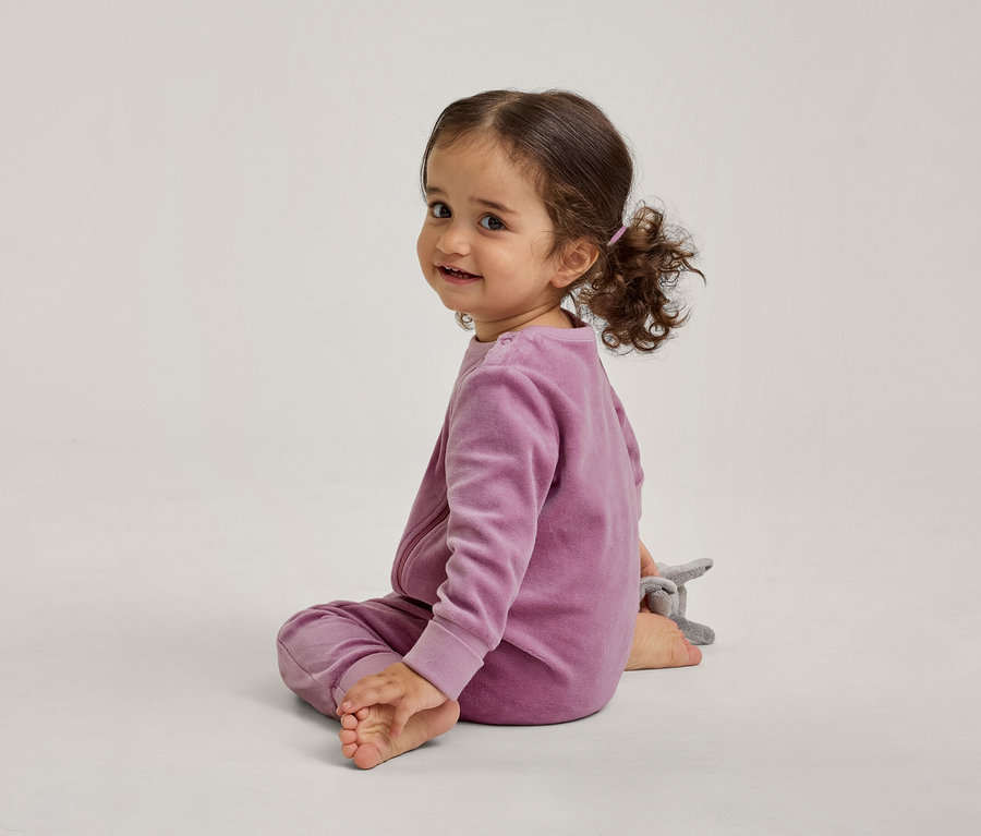 Un tout-petit aux cheveux bouclés foncés, vêtu d'un haut à manches longues et d'un pantalon en velours violet assorti, est assis sur un sol clair. Il regarde par-dessus son épaule gauche, souriant à l'appareil photo, avec un petit animal en peluche gris à côté de lui.