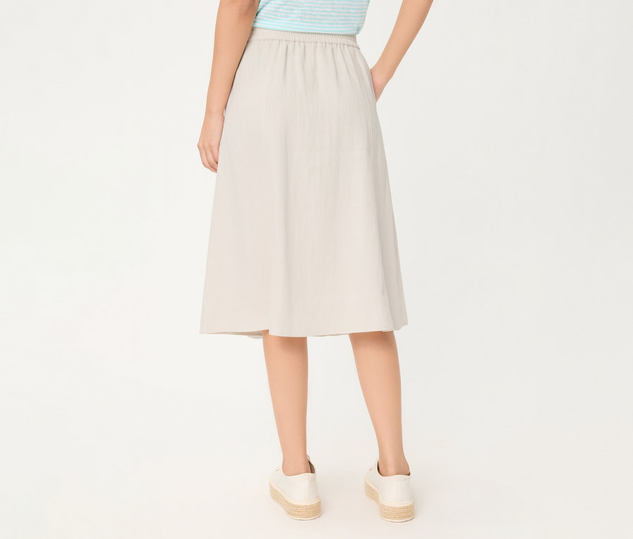 Une personne de dos, portant une jupe en lin Olsen beige à taille élastique, un haut rayé bleu et blanc et des espadrilles blanches. Les mains sont dans les poches.