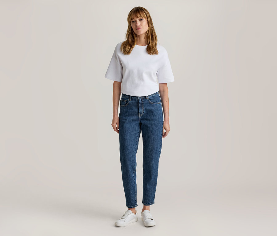 Femme debout, portant un t-shirt blanc à manches courtes, un jean bleu et des baskets blanches.