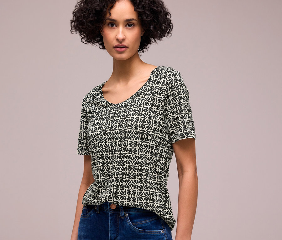Une femme aux cheveux bouclés foncés porte un T-shirt Street One à manches courtes avec un motif de feuilles noires et vert clair et un jean bleu.