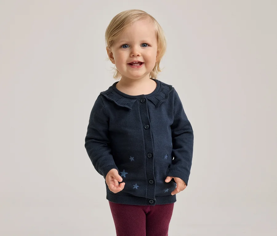 Un enfant souriant aux cheveux blonds et aux yeux bleus porte un cardigan bleu marine à col volanté et motifs étoiles, ainsi qu'un legging bordeaux.