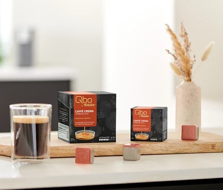 Un verre de café est posé sur une planche en bois, à côté de deux boîtes noires de capsules Qbo Caffè TERRENO ALTO, une de 27 et une de 8 capsules. Deux capsules Qbo individuelles, une brun-rouge et une grise, sont devant les boîtes. Un vase avec de l'herbe séchée est à droite, et un bol de biscuits est à gauche.