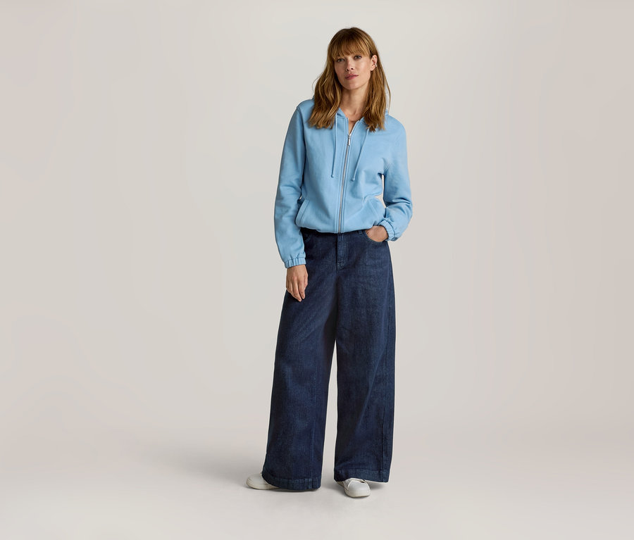 Femme aux longs cheveux châtains clairs portant une veste en sweat zippée bleu clair et un jean large bleu foncé, une main dans la poche.