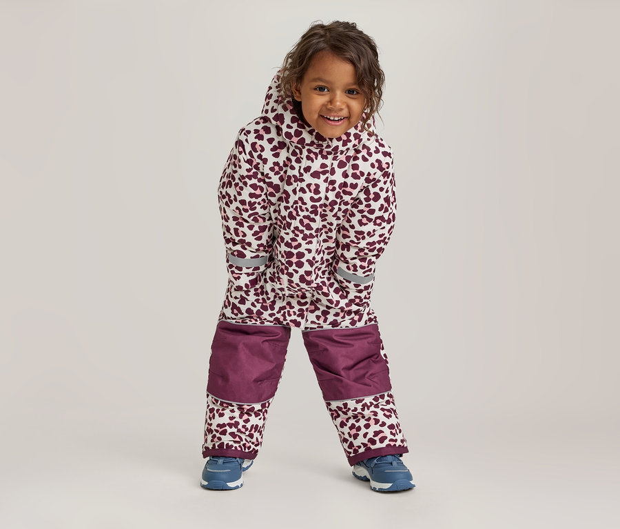 Un enfant souriant porte une combinaison de neige blanche et bordeaux à imprimé léopard, avec des empiècements bordeaux unis aux genoux et des baskets bleues.