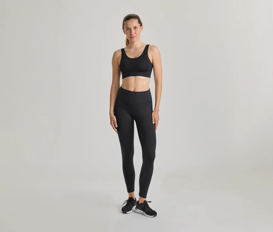 Femme debout, portant un soutien-gorge de sport noir et un legging noir. Baskets noires et blanches, cheveux attachés en arrière. Fond clair et uni.