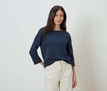Une femme aux longs cheveux bruns porte un chemisier bleu marine à pois blancs et un pantalon beige.