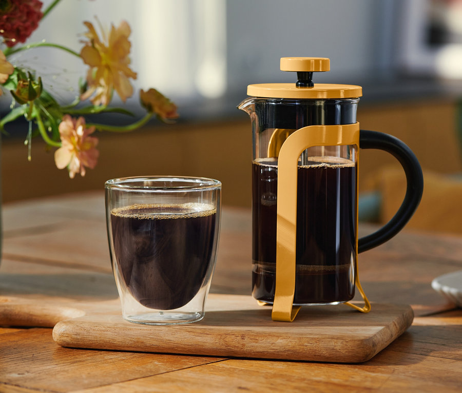 Un verre à double paroi de café noir et une cafetière à piston jaune remplie de café sont posés sur une planche en bois. Un vase de fleurs et une assiette avec un biscuit sont en arrière-plan.