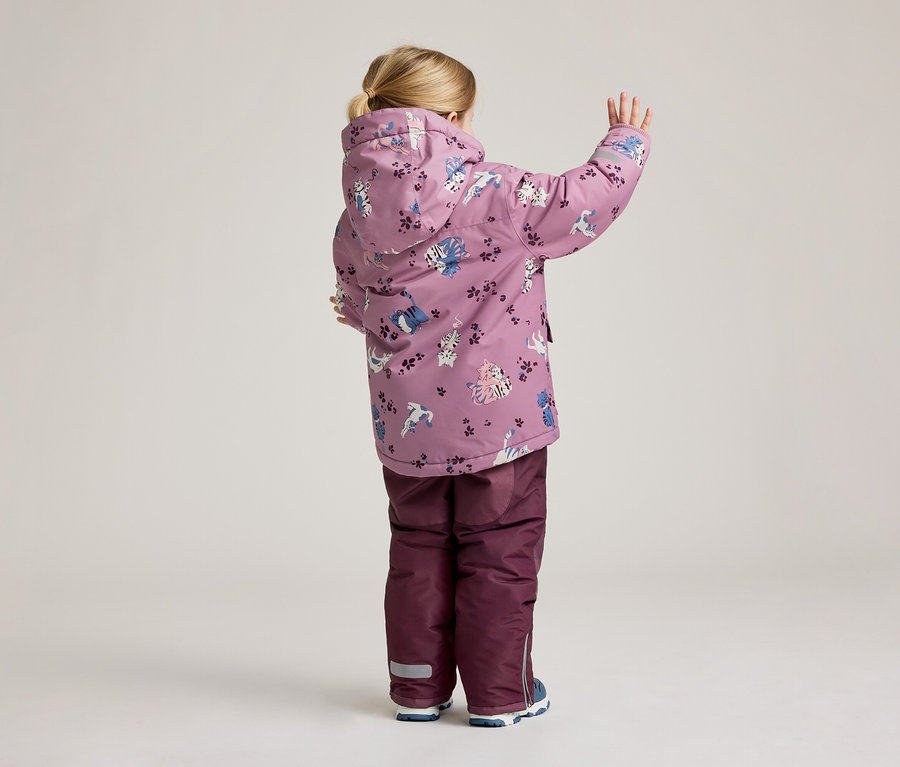 Un enfant vu de dos portant une veste lilas à motif de chats et un pantalon lilas, levant une main.