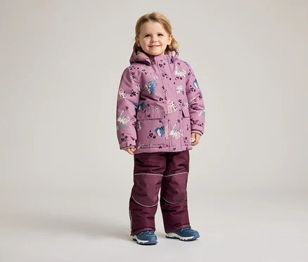 Une petite fille se tient sur un fond clair, vêtue d'un manteau d'hiver violet à motif de chat et d'un pantalon assorti.
