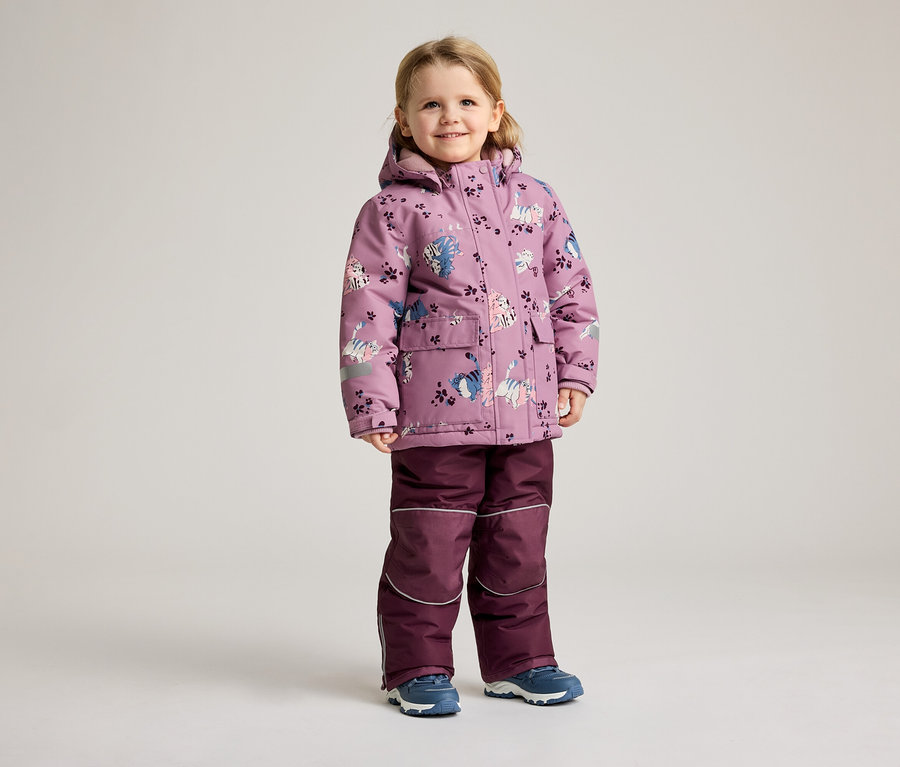 Une petite fille se tient sur un fond clair, vêtue d'un manteau d'hiver violet à motif de chat et d'un pantalon assorti.