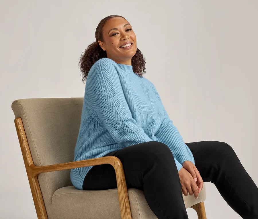 Femme souriante aux cheveux bouclés assise dans un fauteuil, portant un pull bleu clair et un pantalon en bengaline noir.