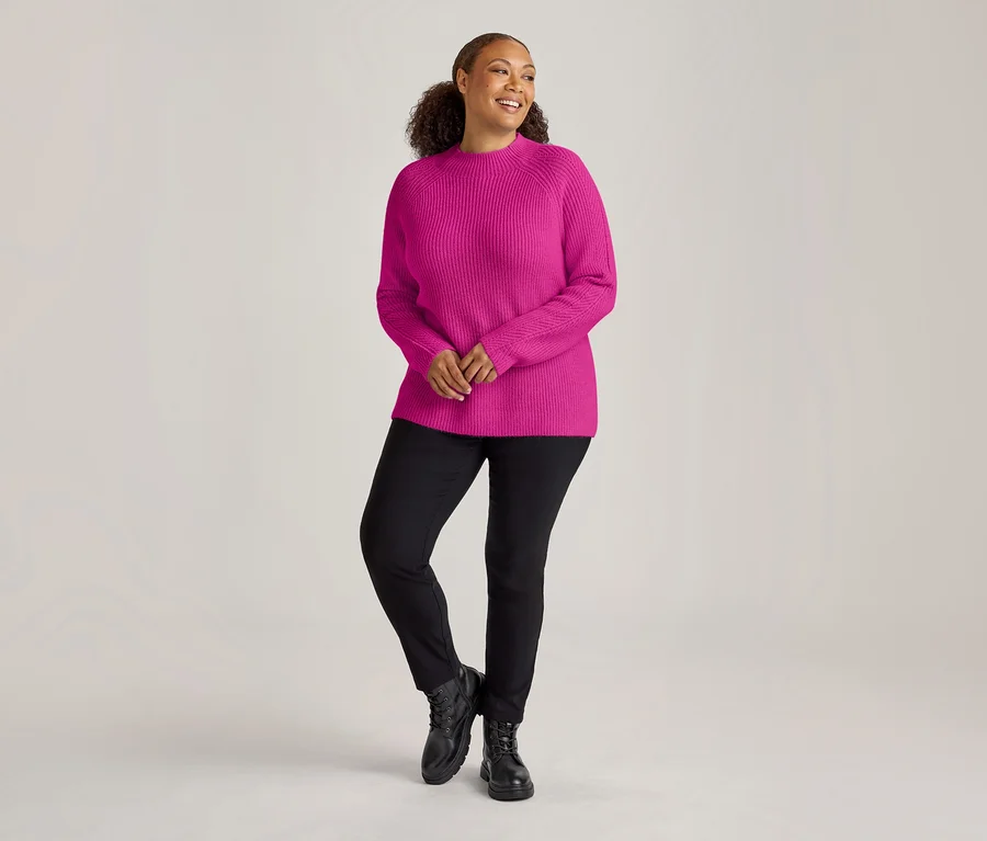 Femme portant un pull côtelé rose vif, un pantalon en bengaline noir et des bottes noires, souriant et regardant sur le côté.