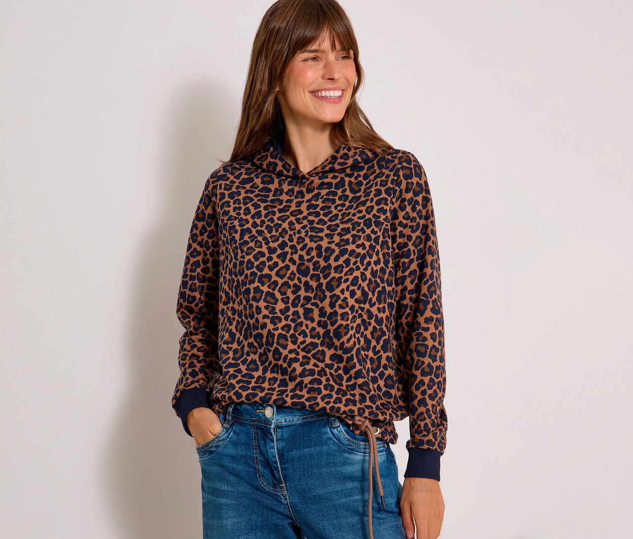 Femme souriante portant un sweatshirt CECIL à imprimé léopard marron et bleu foncé avec capuche et un jean bleu, mains dans les poches.