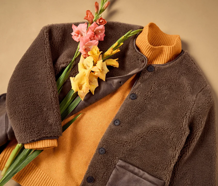 Un cardigan mérinos marron moelleux est posé ouvert sur un pull côtelé orange, avec des glaïeuls roses et jaunes par-dessus.