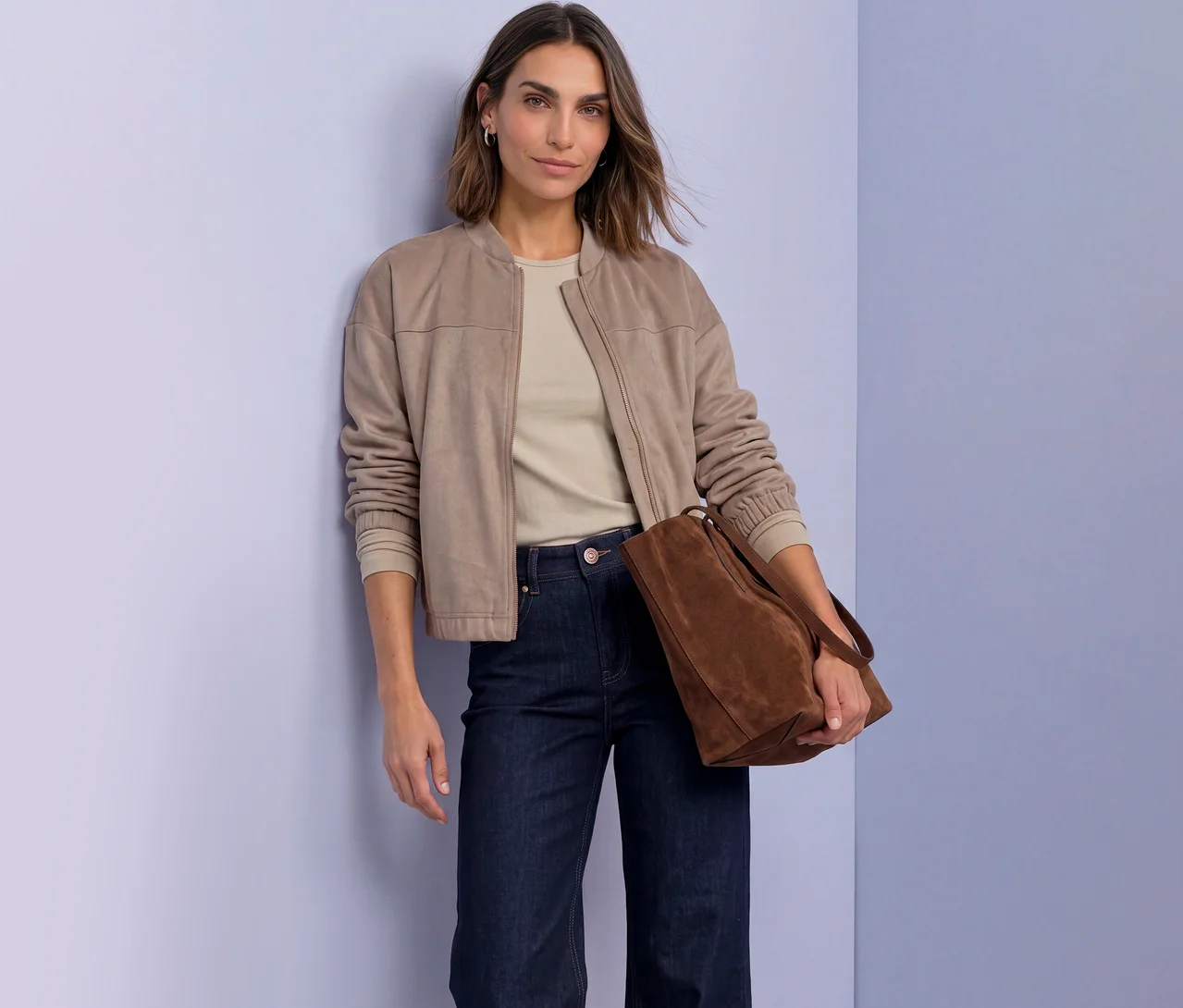 Femme en veste Street One beige, haut clair et jean foncé, tenant un sac cabas marron, se tient devant un mur violet, regardant l'appareil photo.
