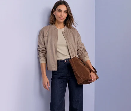 Femme en veste Street One beige, haut clair et jean foncé, tenant un sac cabas marron, se tient devant un mur violet, regardant l'appareil photo.