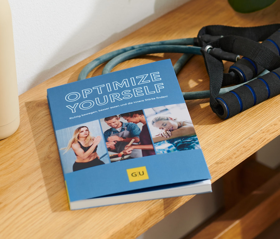 Sur un banc en bois se trouvent le livre « Optimize Yourself », une bouteille d'eau beige et un expandeur.
