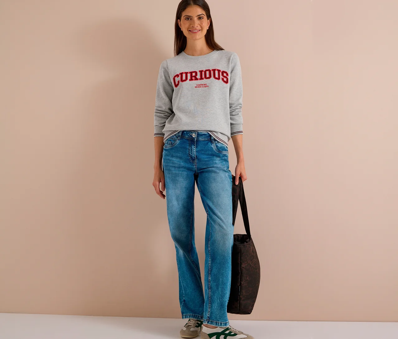 Une femme souriante est debout, vêtue d'un sweatshirt CECIL gris avec l'inscription rouge "CURIOUS", d'un jean bleu et de baskets beiges, tenant un sac foncé.