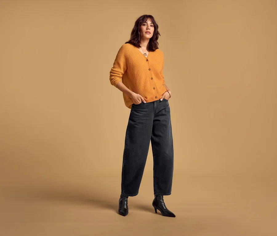 Une femme porte un Merino-Cardigan orange et un Barrel Jeans noir, les mains dans les poches.