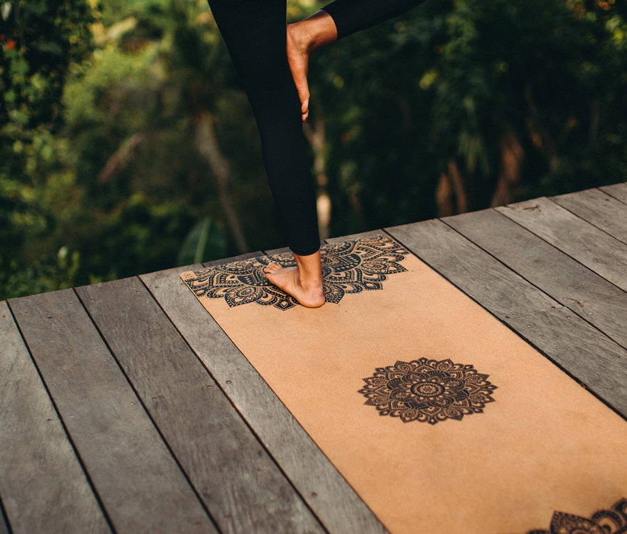 Une femme se tient sur une jambe sur un tapis de yoga avec un motif de mandala sur une terrasse en bois.