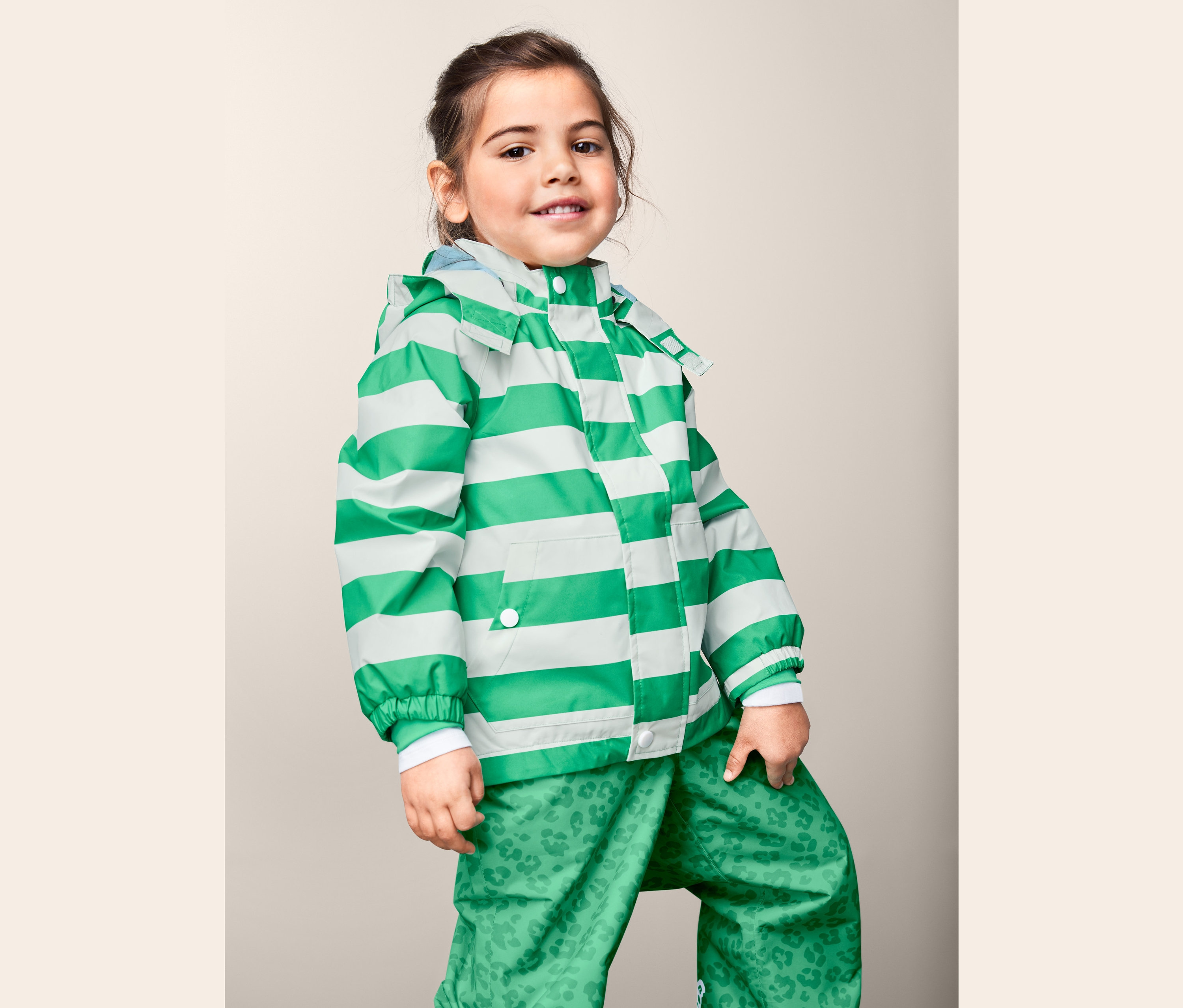 Fillette posant en veste de pluie en matériaux recyclés à rayures vertes et blanches et pantalon vert imprimé.