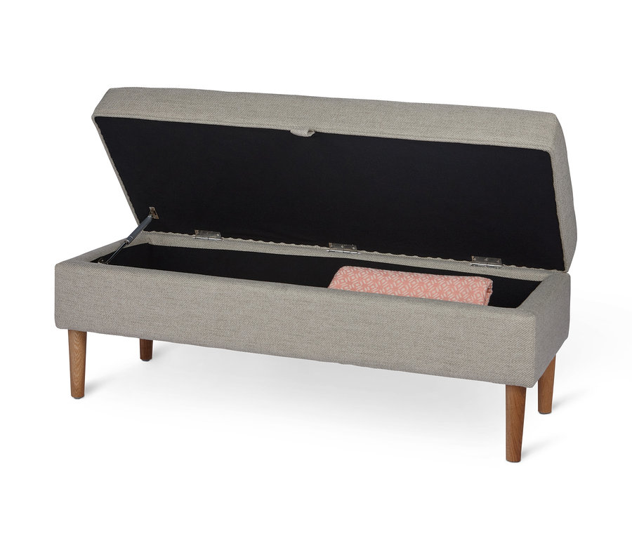 Un banc rembourré avec espace de rangement ouvert, recouvert de beige, repose sur quatre pieds en bois. Une couverture à motifs géométriques se trouve à l'intérieur.
