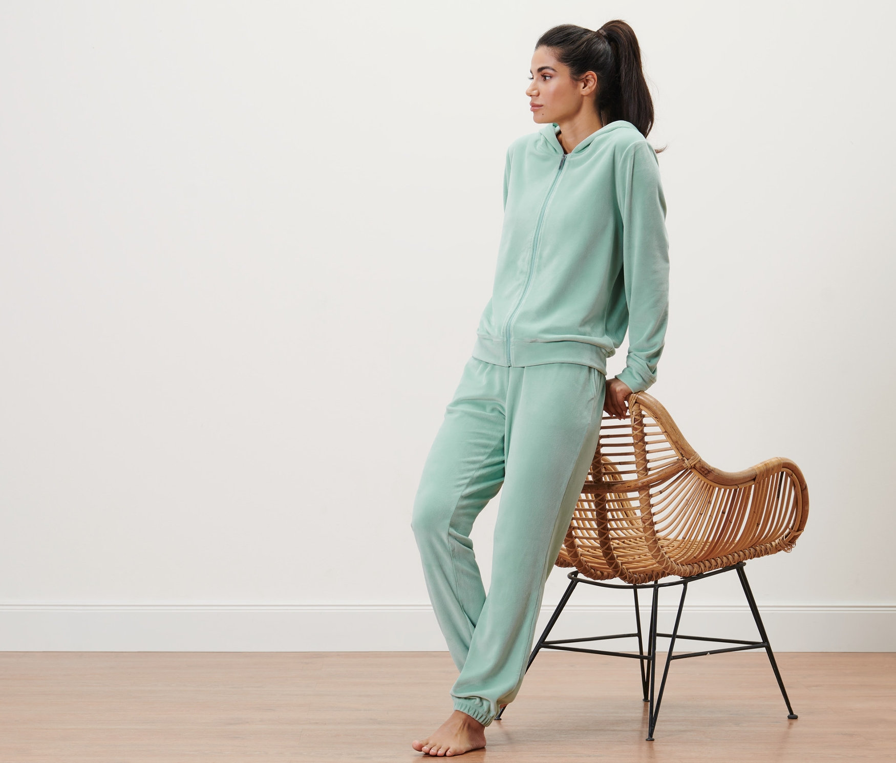 Femme portant un survêtement d’intérieur en velours ras couleur sauge, appuyée contre un fauteuil en osier.