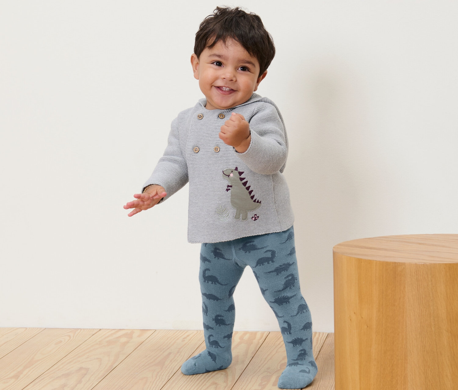 Un jeune enfant souriant se tient debout et porte des collants bleus à motifs de dinosaures.