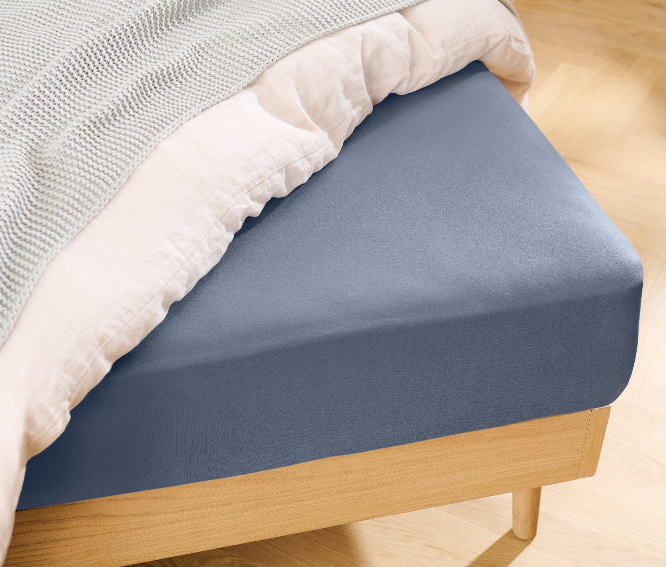 Drap-housse en jersey bleu sur un lit avec une couverture blanche et un couvre-lit tricoté gris.