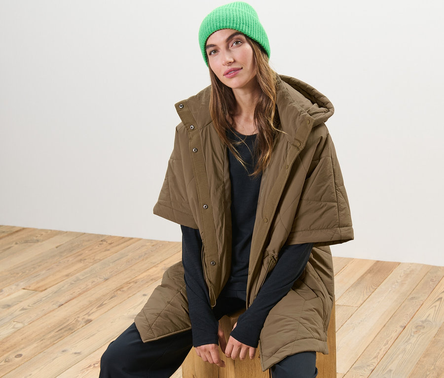 Femme assise sur un tabouret en bois portant un poncho matelassé à capuche et un bonnet vert.