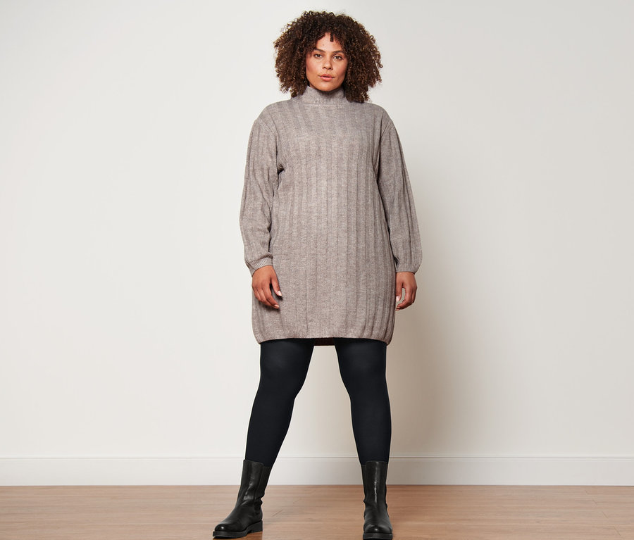 Une femme debout porte une robe en maille côtelée avec col montant, 2 paires de collants opaques noirs et des bottines en cuir.