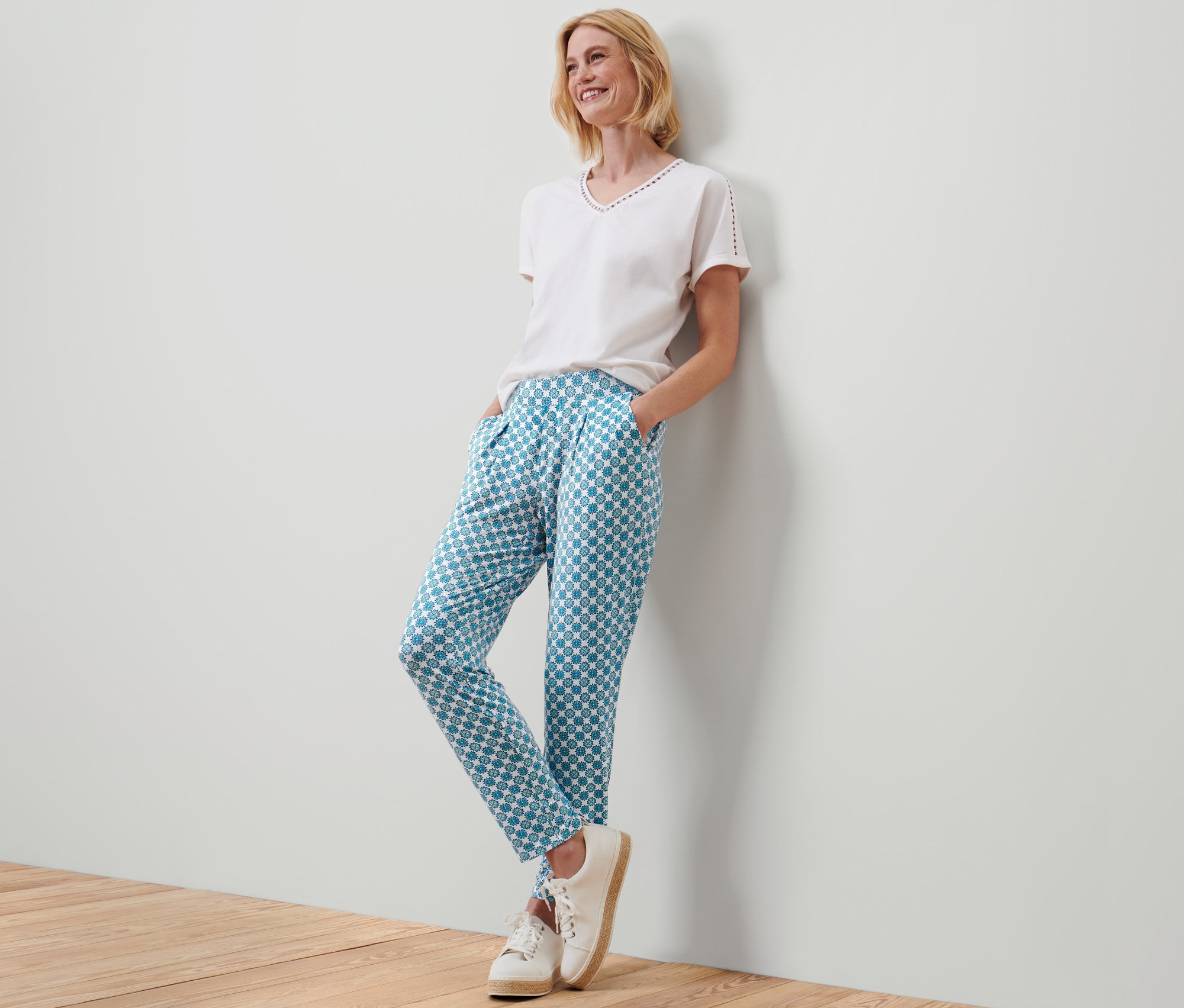 Une femme souriante appuyée contre un mur porte un pantalon en jersey avec motif intégral.