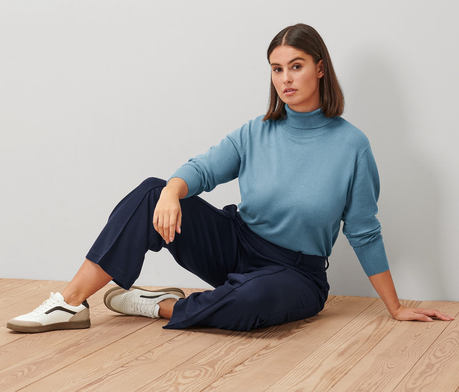 Une femme est assise sur un plancher en bois, portant un pull à col roulé en maille fine bleu fumée, un pantalon en jersey bleu foncé avec nervure et des baskets en cuir.