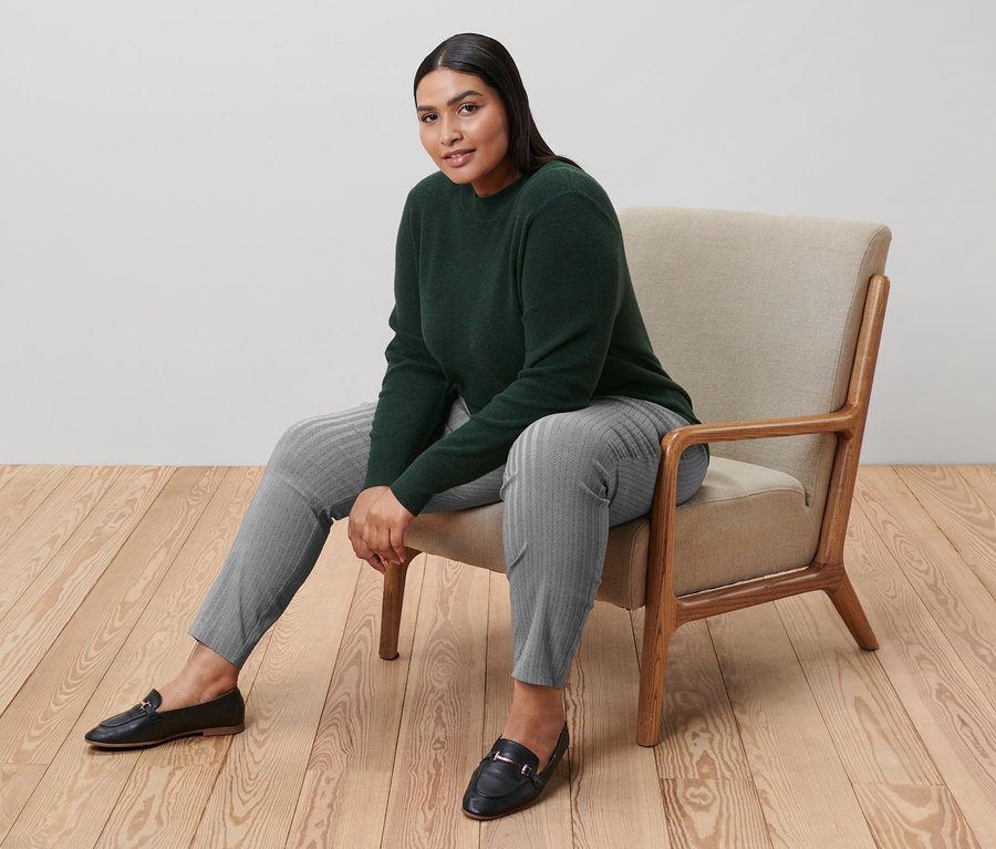 Femme assise dans un fauteuil portant un pull en maille fine vert et un pantalon stretch imprimé gris.