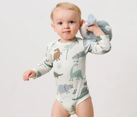 Un bébé tient une peluche et porte un 3	bodys manches longues, dinosaure.