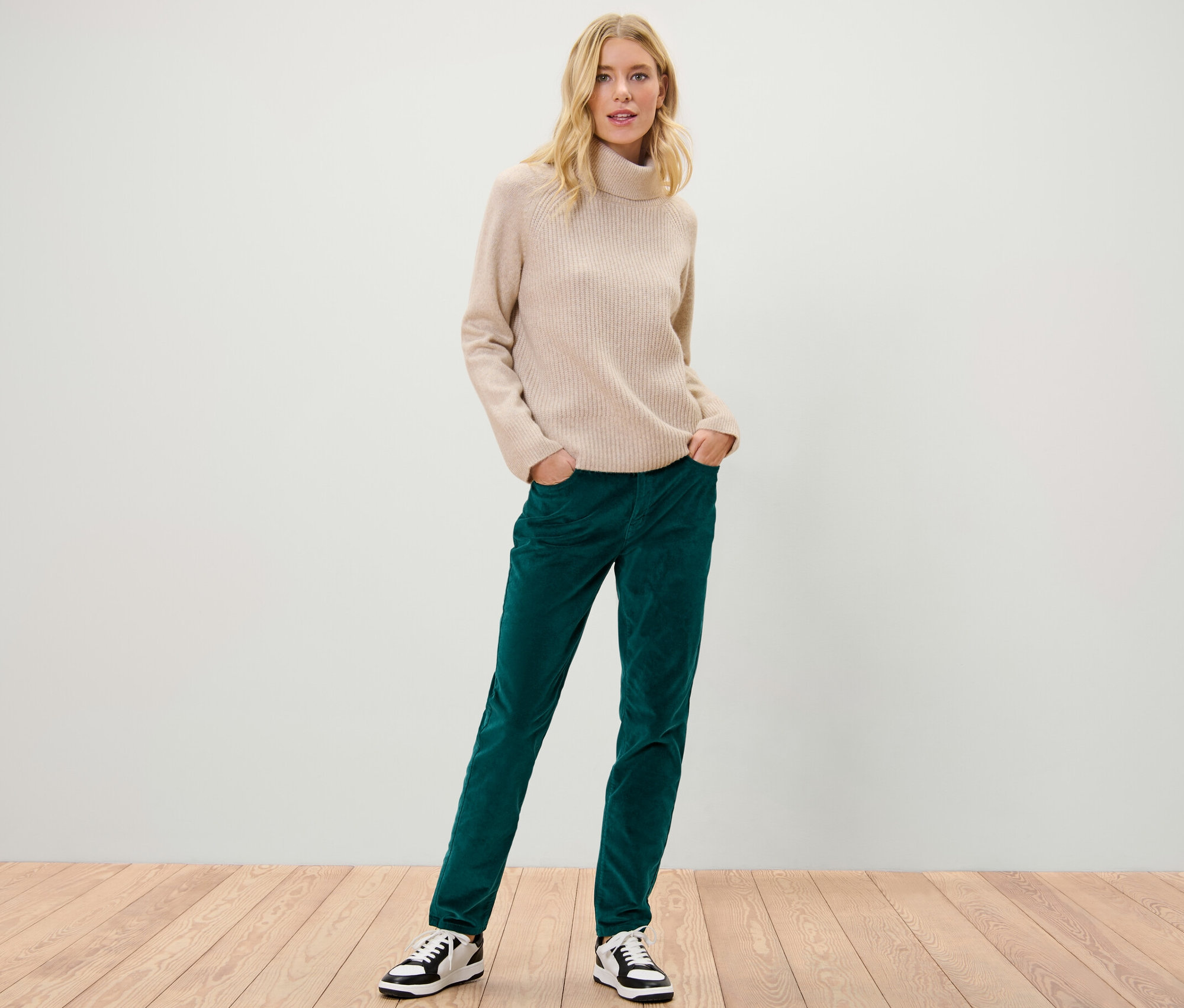 Une femme blonde pose en pantalon en velours pétrole et pull à col roulé beige.