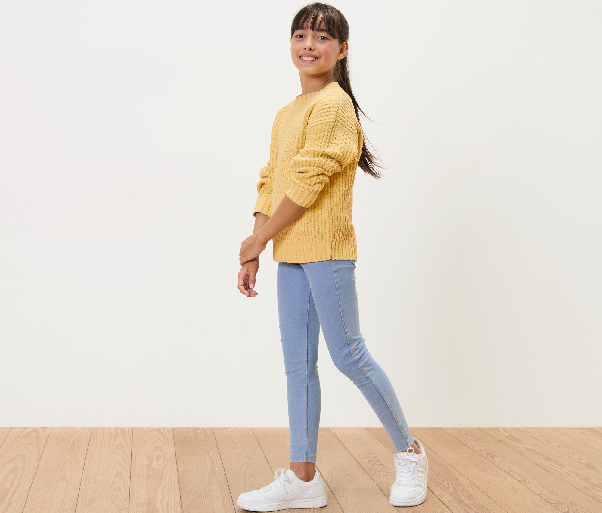 Une fille pose en pull chenille jaune colza avec un jean bleu clair et des baskets blanches.