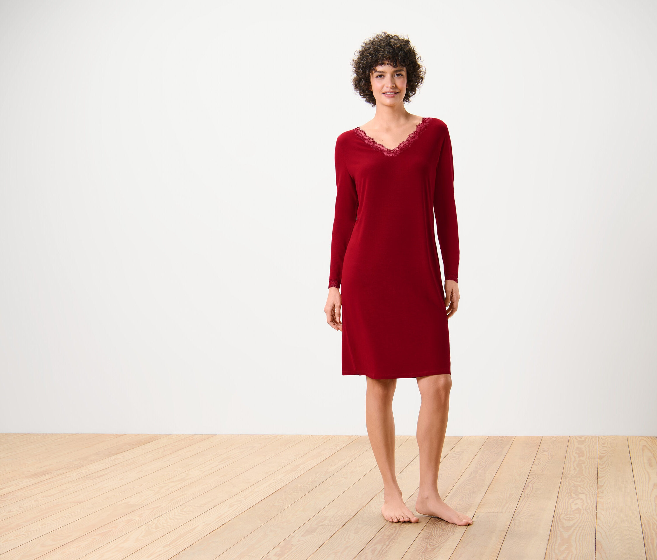 Une femme en chemise de nuit rouge à manches longues avec de la dentelle à l'encolure se tient sur un plancher en bois devant un mur blanc.