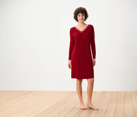 Une femme en chemise de nuit rouge à manches longues avec de la dentelle à l'encolure se tient sur un plancher en bois devant un mur blanc.